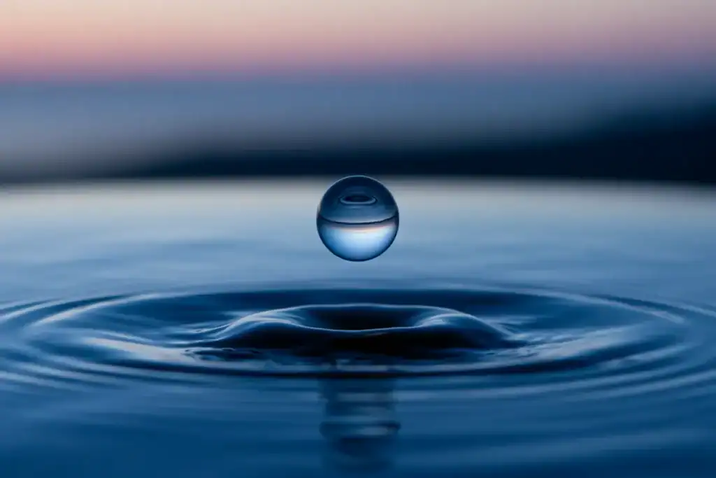 A close-up of a water droplet captured just before it touches the surface, where soft ripples are already forming. The background is out of focus and ethereal. Represents chiron healing in motion, impact, and resonance.