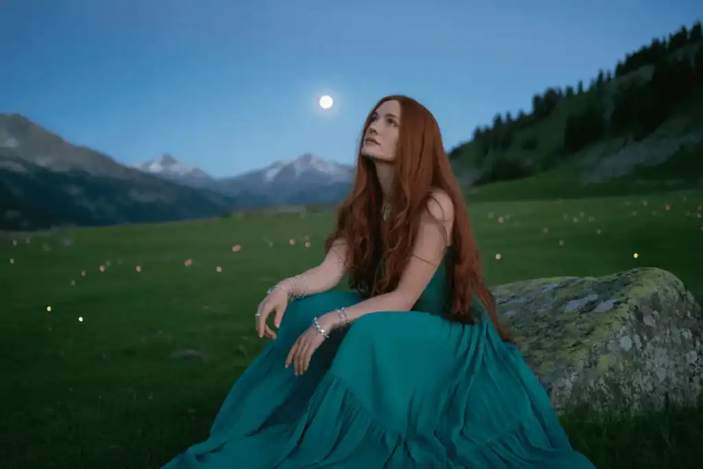 Red-haired woman in a flowing green dress sits on a rock under the full moon, gazing at the sky in a peaceful mountain landscape.