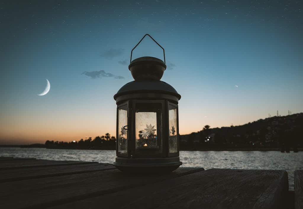 Serene evening scene featuring a lantern on a dock by the water under a crescent moon and starry sky.